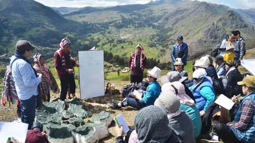 A group of people sit on the floor and listen to two people presenting in front of a scenic mountain backdrop.