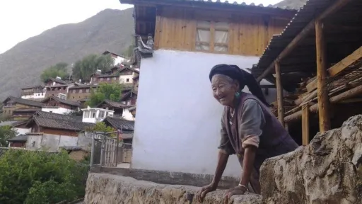 An elderly woman leans over a stone wall with traditional terrace houses behind her.
